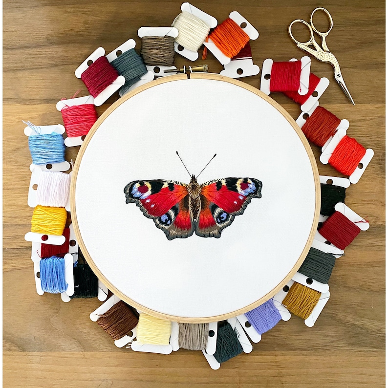 May include: A white embroidery hoop with a colorful peacock butterfly design. The hoop is surrounded by colorful embroidery floss spools.