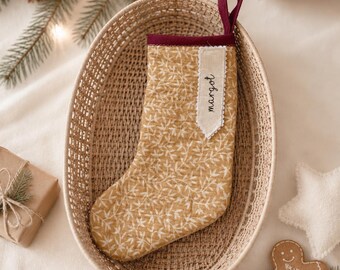 May include: A quilted Christmas stocking in mustard yellow with a white leaf pattern, featuring a dark burgundy cuff and a hanging loop. A cream-colored fabric tag is sewn onto the cuff, displaying the name "margot" in black cursive script.
