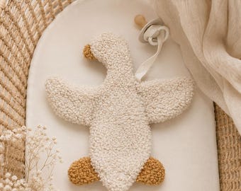 May include: A cream-colored, textured, sherpa-style fabric goose toy with a tan beak and tan feet, accompanied by a white pacifier attached to a white fabric strap. The goose has outstretched wings and a soft, plush appearance.