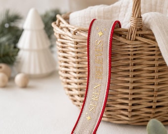 May include: A pacifier clip featuring a beige fabric strap with red trim, gold embroidered text reading "SEREN BELIEVES" flanked by two small gold stars, and a round, light-colored wooden pacifier attachment with three ventilation holes.