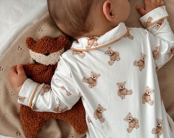 May include: A baby wearing a white onesie with a teddy bear pattern, trimmed with tan piping. The baby is snuggled with a brown plush fox toy on a tan blanket. The scene is captured from above.