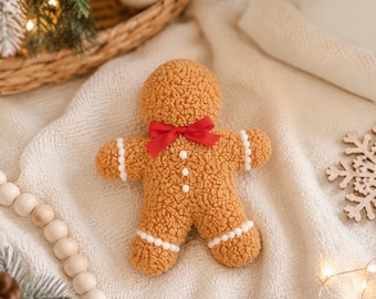 May include: A tan, textured, sherpa-style gingerbread man plush toy featuring a bright red ribbon bow at the neck, three small white pom-pom buttons down the center, and white pom-pom trim accents on the arms and legs.