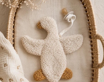 May include: A cream-colored, textured goose-shaped plush toy with a mustard yellow beak and feet. A white pacifier with a matching white fabric clip is attached to the goose. The toy is resting inside an oval-shaped woven wicker basket.