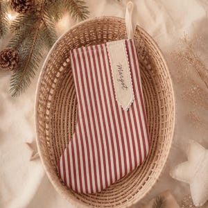May include: A Christmas stocking featuring a red and white vertical striped pattern. The stocking includes a cream-coloured fabric tag sewn onto the front with the name Margot written in black script and a fabric loop at the top for hanging.