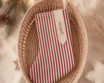 May include: A Christmas stocking featuring a red and white vertical striped pattern. The stocking includes a cream-colored fabric tag sewn onto the front with the name Margot written in black script and a fabric loop at the top for hanging.