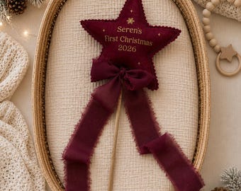 May include: A burgundy felt star ornament on a wooden stick, featuring the embroidered text "Seren's First Christmas 2026" in gold. The star is adorned with a matching burgundy frayed ribbon bow and small gold star accents, creating a festive decoration.