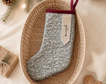May include: A Christmas stocking featuring a light blue fabric with a white botanical leaf pattern, a dark red trim, and a hanging loop. A beige fabric tag is attached to the top, displaying the name "margot" in black cursive script.