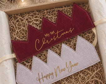 May include: Two felt crowns in a gift box. The top crown is deep red with the gold text My 1st Christmas. The bottom crown is light gray with the gold text Happy New Year. Both crowns feature a jagged top edge and are tied with cream ribbons.