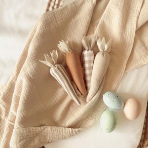 May include: A selection of fabric carrots in different colours and patterns, including striped and checkered designs, with green fabric tops. The carrots are arranged on a beige cloth with three speckled Easter eggs in pastel colours.