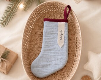 May include: A light blue fabric Christmas stocking with a dark red trim and a hanging loop. A cream-colored fabric tag is sewn onto the front, featuring the name "margot" embroidered in black cursive script with a decorative scalloped edge.