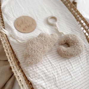 May include: A baby mobile and wooden sign in a wicker bassinet. The mobile has a fluffy cloud and crescent moon shape. The wooden sign reads "hello world". The bassinet has a striped, cream-coloured sheet.