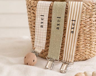 May include: Three baby pacifier clips with the name "axel" printed on them. The clips have different patterns: a beige gingham, a textured olive green, and a beige and white stripe. A wooden teether and a woven basket are in the background.