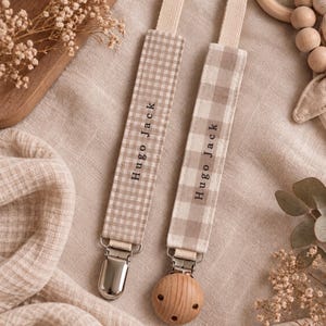 Peut inclure: Deux attaches-sucettes en tissu avec un motif vichy beige et blanc. Chaque sangle est brodée du nom Hugo Jack en fil noir. Une attache possède une pince en métal argenté, tandis que l'autre présente une pince ronde en bois clair.