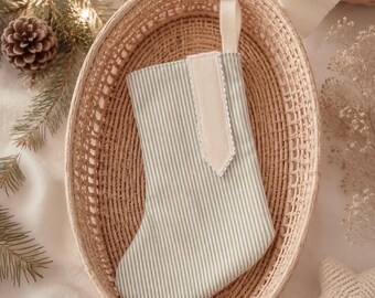 May include: A Christmas stocking featuring a light green and white pinstripe pattern, accented with a solid cream-colored vertical tab and a matching cream-colored hanging loop at the top, finished with a delicate scalloped edge trim.