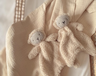 May include: Two plush, cream-colored lamb security blankets rest on a soft, beige blanket. The lambs have gray faces and small, embroidered eyes. The blanket is draped over a white surface, creating a cozy and comforting scene.