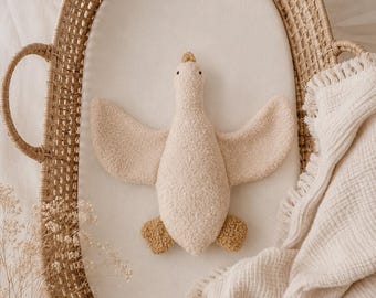 May include: A plush goose toy with a cream-colored textured body, small dark eyes, and mustard-yellow feet, resting inside an oval-shaped woven wicker basket with handles, accompanied by a cream-colored muslin blanket with fringed edges.