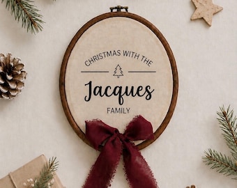 May include: An oval wooden embroidery hoop containing cream-colored fabric with the black printed text CHRISTMAS WITH THE, a small tree icon, the name Jacques, and the word FAMILY. A dark red frayed ribbon bow is attached to the bottom.