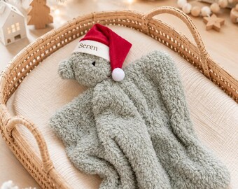 May include: A sage green plush security blanket featuring a stuffed animal head wearing a red and white Santa hat with the name Seren embroidered on the brim. The blanket is made of a soft, textured, fuzzy material with a square-shaped body.