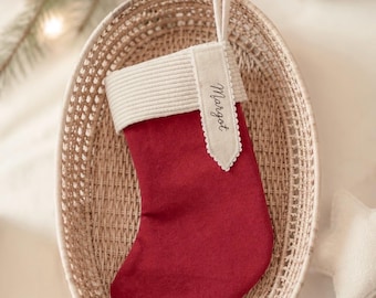 May include: A deep red Christmas stocking featuring a cream-colored ribbed cuff and a matching cream fabric tag with the name Margot embroidered in black script. The stocking includes a small fabric loop for hanging at the top of the cuff.