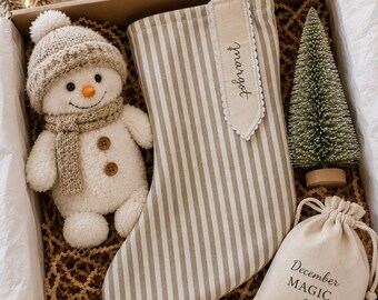 May include: A gift box containing a beige and white striped Christmas stocking with the name Margot embroidered on a tag, a plush snowman wearing a knit hat and scarf, a small artificial pine tree, a drawstring bag labeled December MAGIC, and a candy cane.