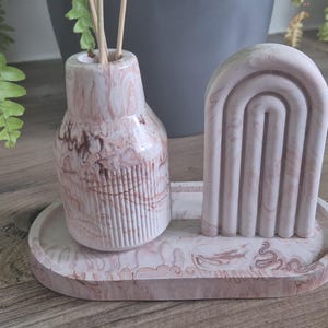 May include: A decorative set with a marble-effect tray, a vase with reeds, and an arched sculpture. The items are in shades of pink and brown. The oval tray holds the vase and sculpture. The vase has vertical lines.