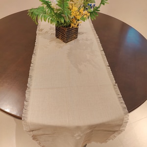 May include: A beige table runner with a fringed edge. The runner is on a dark brown round table with a bouquet of white and yellow flowers in a brown planter.