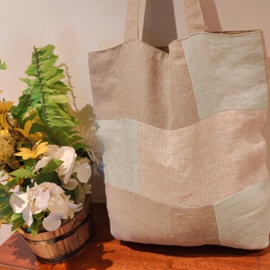 May include: A beige and light blue patchwork tote bag with a long handle. The bag is made of a soft, natural fabric and has a relaxed, casual style.