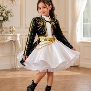 May include: A black sequined bolero jacket with gold trim worn over a white dress featuring a high neckline and a full skirt. The outfit includes a gold coin belt with a draped chain detail and black lace-up boots with a glossy finish.