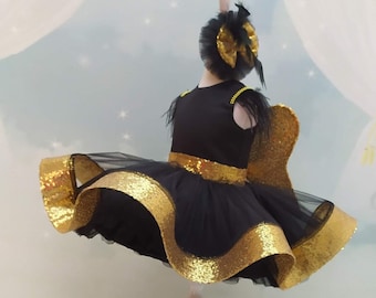 May include: A black tulle dress with gold glitter trim and a matching headband. The dress has a ruffled skirt and is on a white stand. The dress is paired with black shoes with gold glitter butterfly accents.