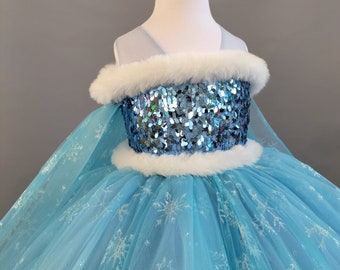 Puede incluir: Un vestido de tul azul claro con ribete de piel blanca y un corpiño de lentejuelas. El vestido está decorado con motivos de copos de nieve.
