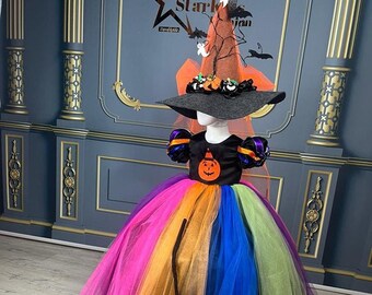 May include: A rainbow tulle dress with a black bodice featuring a pumpkin design. The dress is paired with a black witch hat with orange and brown accents and a black broom.