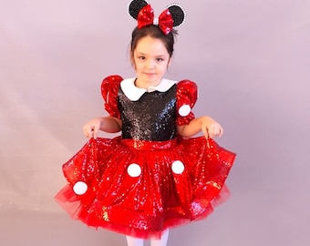 May include: A young girl wearing a red and black sequined dress with white polka dots and a black bow headband with white ears. She is standing on a light gray background.