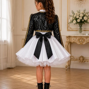 May include: A black sequined bolero jacket with gold trim worn over a white dress featuring a full, pleated skirt and a large black satin bow at the waist. The ensemble is paired with black lace-up boots, creating a formal look with contrasting textures.