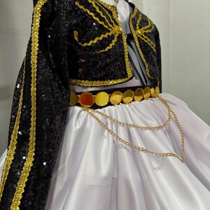 May include: A black sequined bolero jacket with gold trim paired with a white satin skirt. The outfit features a black belt adorned with circular gold metallic discs and a decorative gold chain draped across the front of the white skirt.