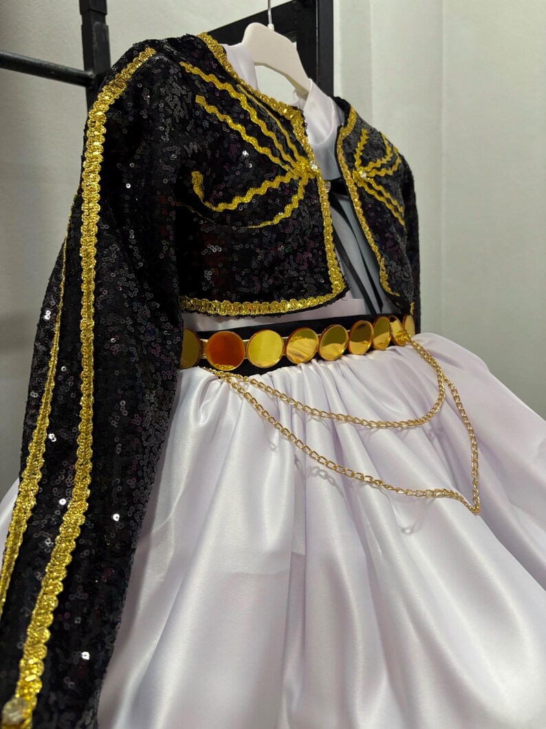 May include: A black sequined bolero jacket with gold trim paired with a white satin skirt. The outfit features a black belt adorned with circular gold metallic discs and a decorative gold chain draped across the front of the white skirt.