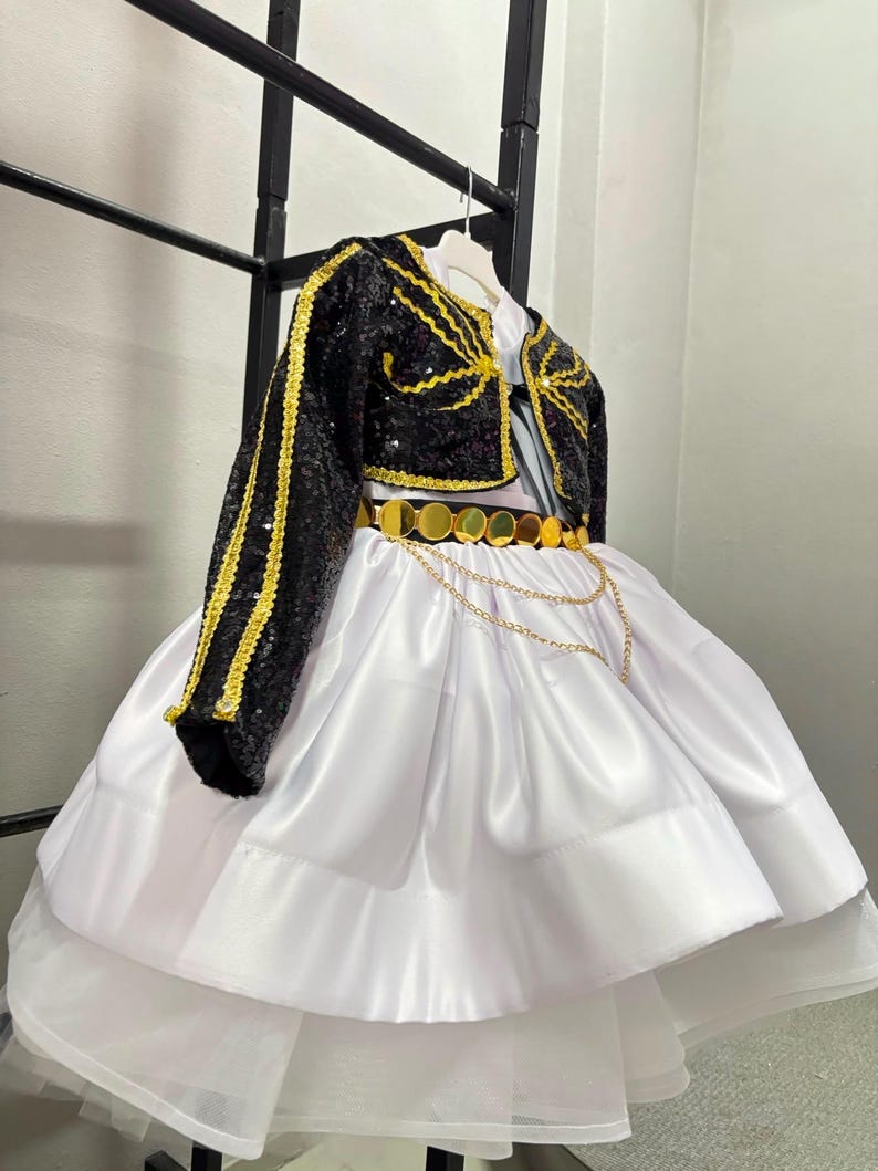 May include: A black sequined bolero jacket with gold trim paired with a white satin dress. The dress features a gold circular disc belt and a layered skirt with a tulle hem. The ensemble is displayed on a white hanger against a black frame.
