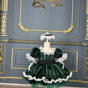 May include: A green and black striped dress with white lace trim and a black sash. The dress is on a white mannequin and has a green tulle underskirt. The dress is for a child.