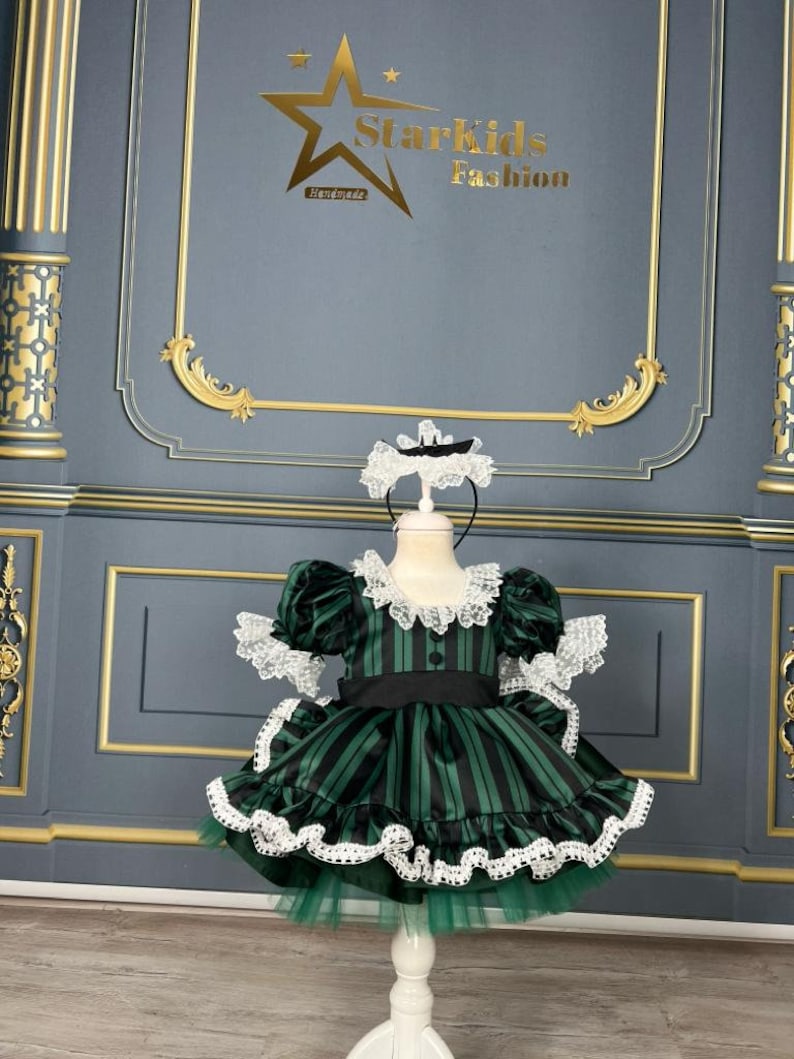 May include: A green and black striped dress with white lace trim and a black sash. The dress is on a white mannequin and has a green tulle underskirt. The dress is for a child.