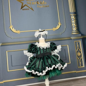 May include: A green and black striped dress with white lace trim and a black bow. The dress is on a white stand against a grey background with gold trim.