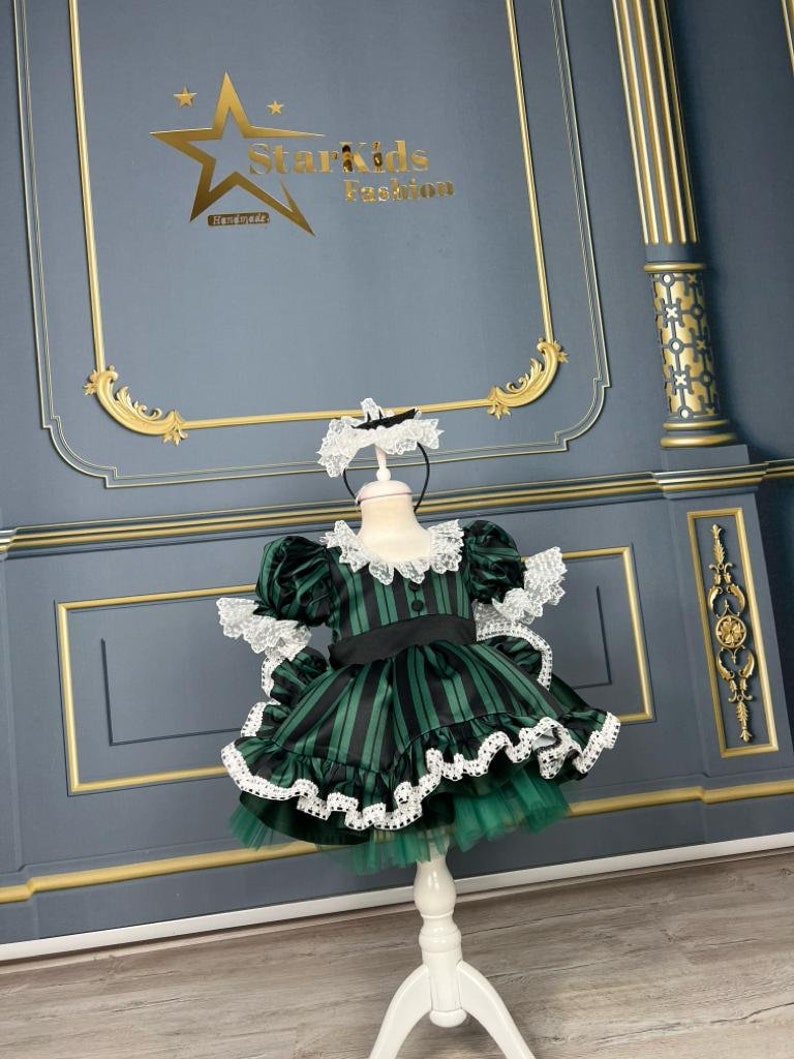 May include: A green and black striped dress with white lace trim and a black bow. The dress is on a white stand against a grey background with gold trim.