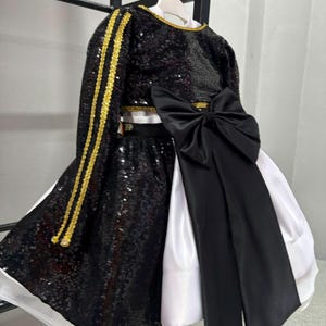 May include: A two-piece outfit featuring a black sequined long-sleeve top and a matching skirt with gold trim accents. The skirt is layered over a white fabric base and is adorned with a large black satin bow at the waist, creating a high-contrast look.
