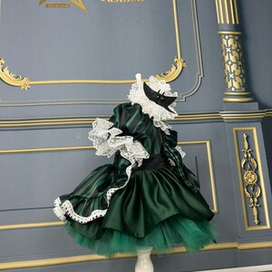 May include: A green and black striped dress with a white lace trim and a black satin sash. The dress has a tulle skirt and a white lace collar. The dress is on a white stand against a grey background. The text "StarKids Fashion" is visible in the top left corner of the image.