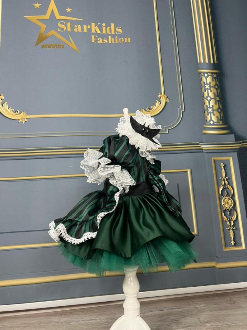 May include: A green and black striped dress with a white lace trim and a black satin sash. The dress has a tulle skirt and a white lace collar. The dress is on a white stand against a grey background. The text "StarKids Fashion" is visible in the top left corner of the image.