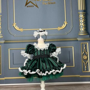 May include: A green and black striped dress with white lace trim and a white bow. The dress is on a white stand against a grey background. The text "Starkids Fashion Handmade" is visible in the top centre of the image.