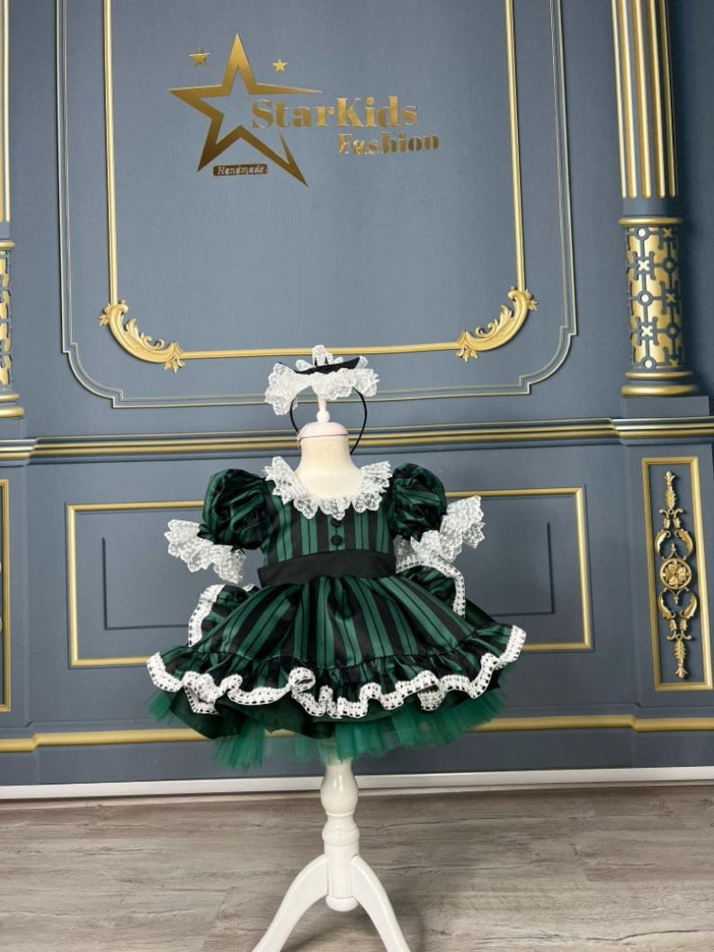 May include: A green and black striped dress with white lace trim and a white bow. The dress is on a white stand against a grey background. The text "Starkids Fashion Handmade" is visible in the top centre of the image.