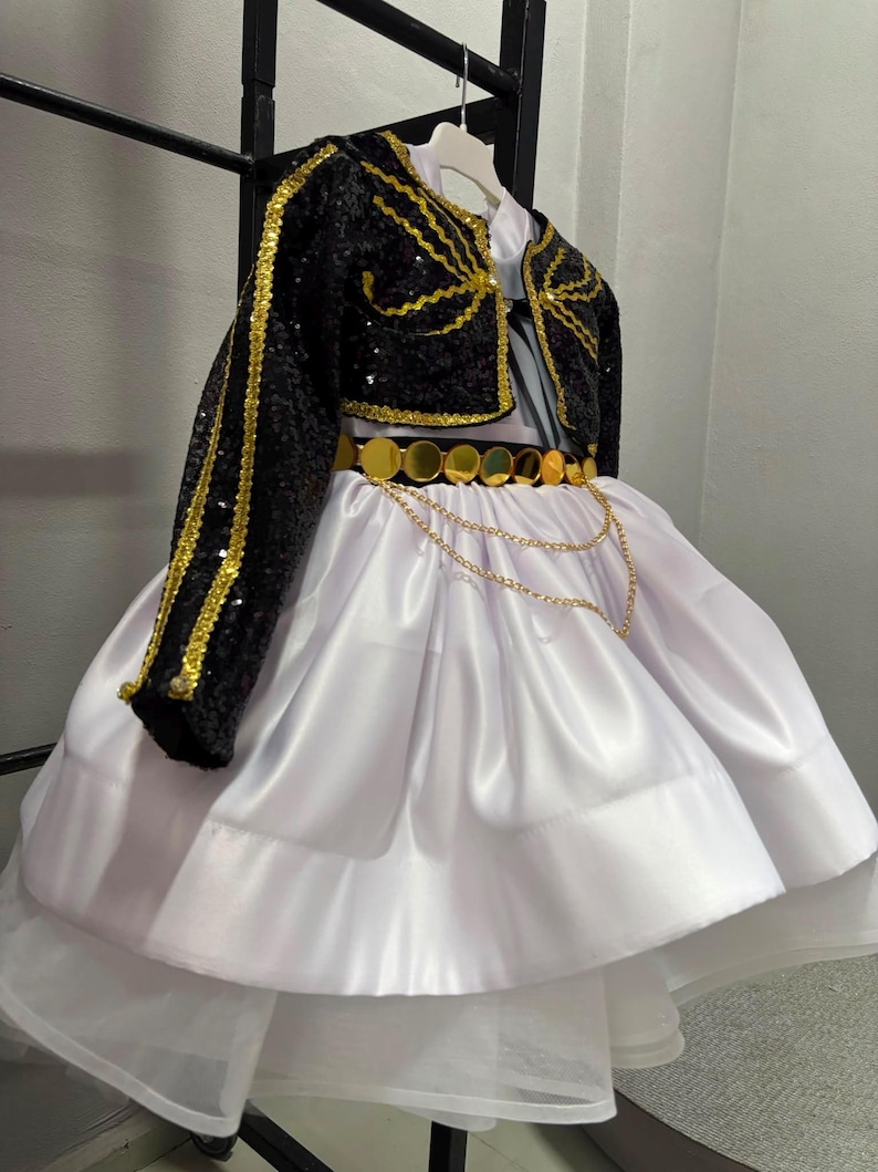 May include: A white satin skirt with a layered tulle hem paired with a black sequined bolero jacket featuring gold trim. The outfit includes a gold metallic disc belt adorned with two draped gold chains, creating a structured and ornate appearance.