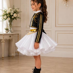 May include: A black sequined bolero jacket with gold trim worn over a white dress with a full, layered skirt. The outfit features a gold coin belt with a chain detail at the waist and is paired with black lace-up boots, creating a structured look.