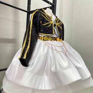 May include: A white dress with a full, layered skirt and a black sequined bolero jacket. The jacket features gold trim and embroidery. A gold belt with circular metallic discs and a draped gold chain accent sits at the waist of the white dress.