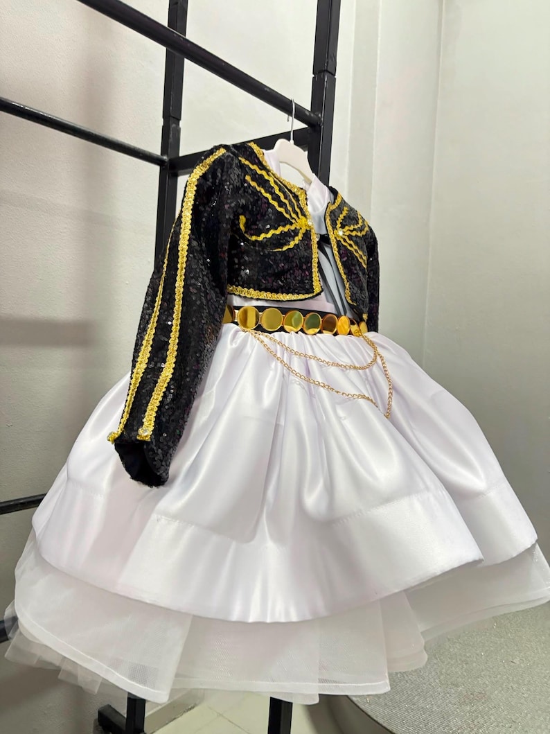 May include: A white dress with a full, layered skirt and a black sequined bolero jacket. The jacket features gold trim and embroidery. A gold belt with circular metallic discs and a draped gold chain accent sits at the waist of the white dress.