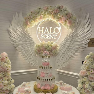 May include: A display for Halo Scent featuring a tiered perfume bottle arrangement on a round white table. The table is decorated with pink handbags, rose petals, and small perfume bottles. The backdrop includes a neon sign with the Halo Scent logo, angel wings, and floral arrangements.
